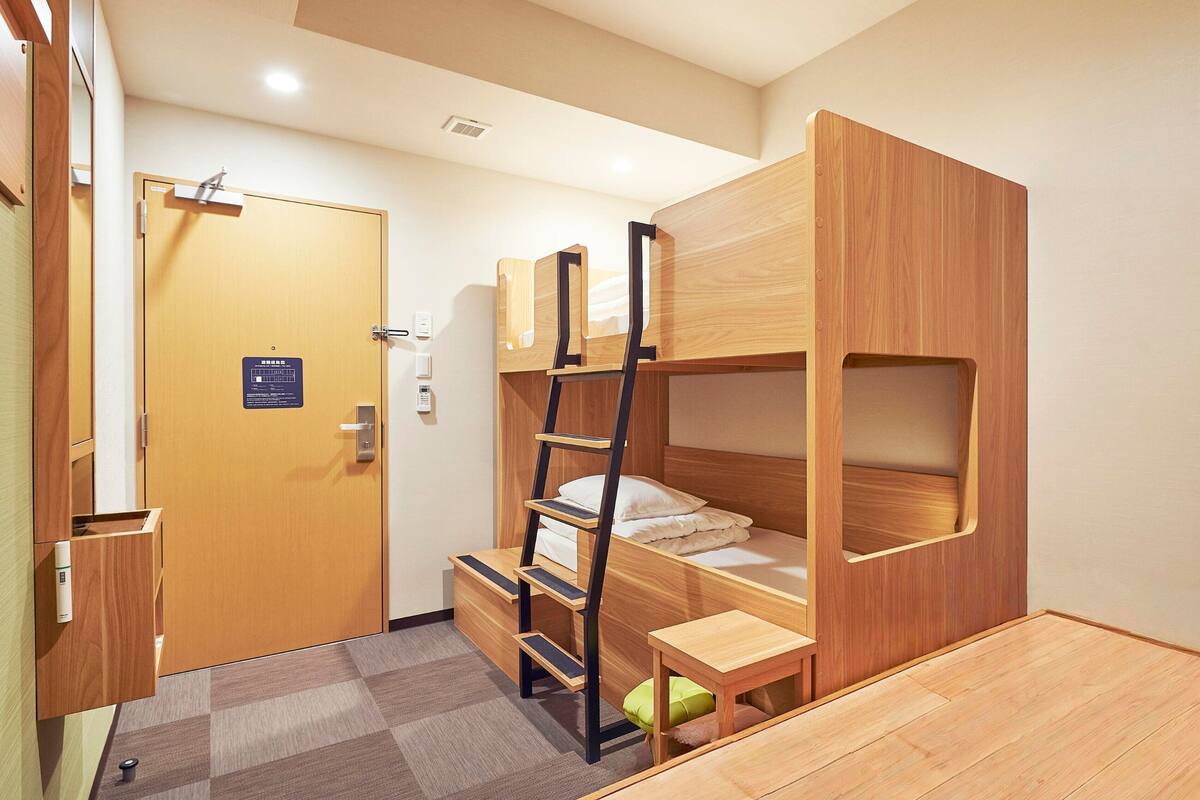 Ein kleines Zimmer mit einem Etagenbett, einem kleinen Schreibtisch und einem Stuhl. An der Wand befinden sich eine Tür mit einem Schild, ein Lichtschalter und ein Thermostat.