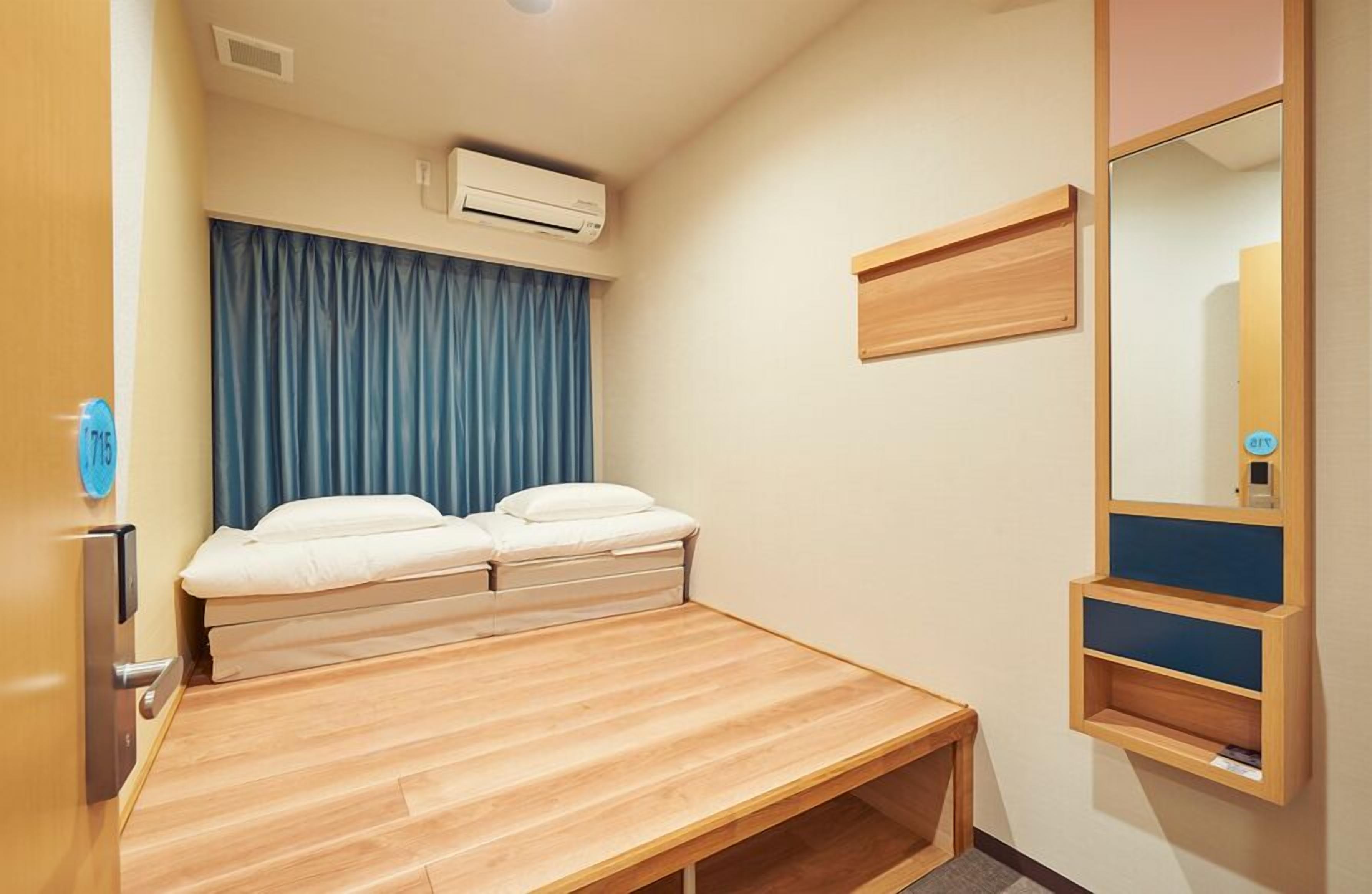 Une petite chambre d’hôtel moderne, équipée d’un lit, d’un bureau en bois, d’une étagère fixée au mur et d’un miroir.