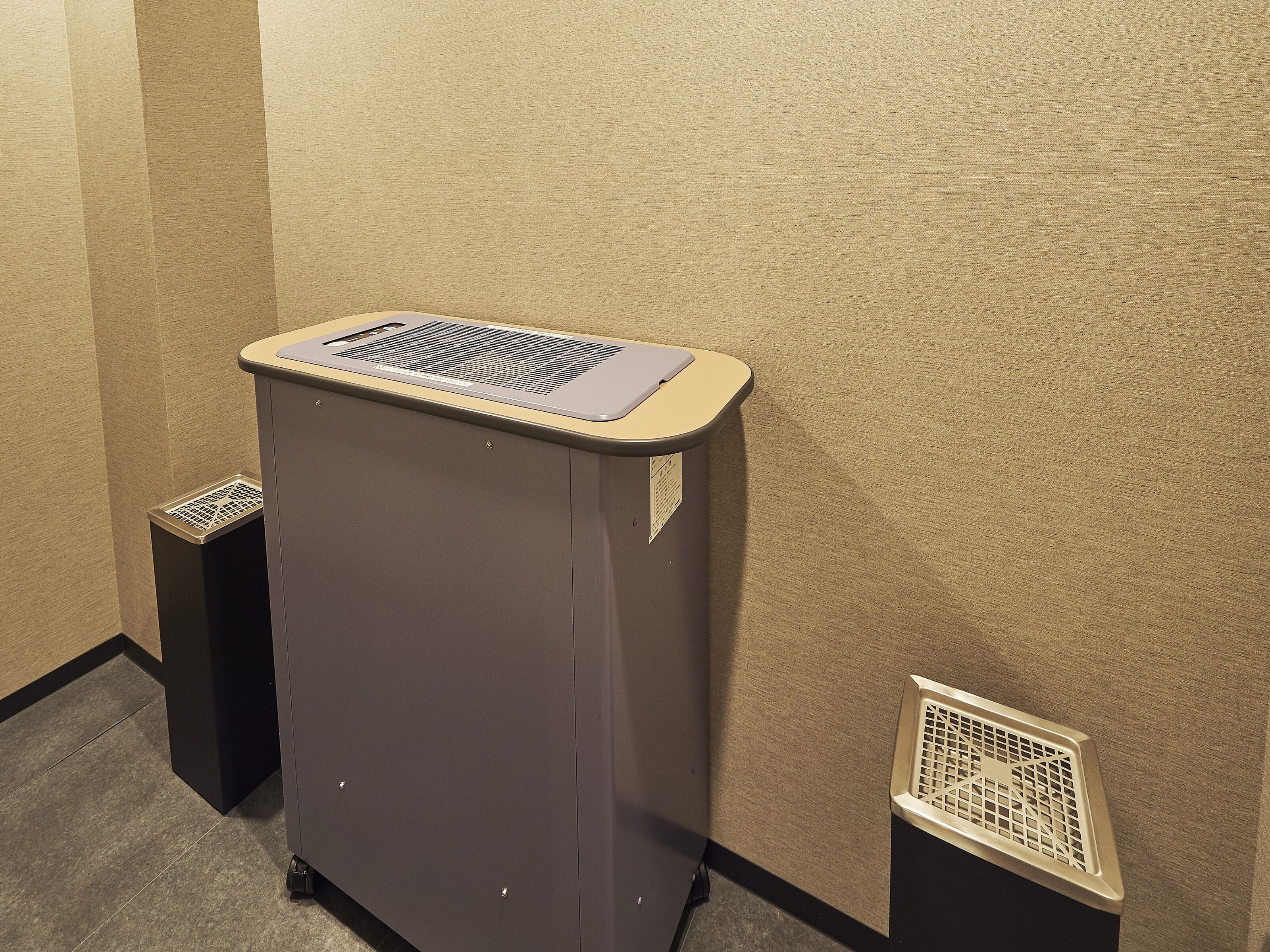 Un petit bureau de couleur sombre, surmonté d’une grille de ventilation, est placé contre un mur beige, près d’une bouche d’aération située au sol.