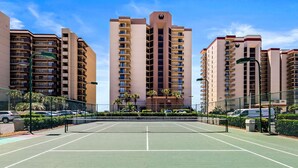 Un court de tennis entouré de grands immeubles dotés de balcons, d’une clôture et de voitures garées.