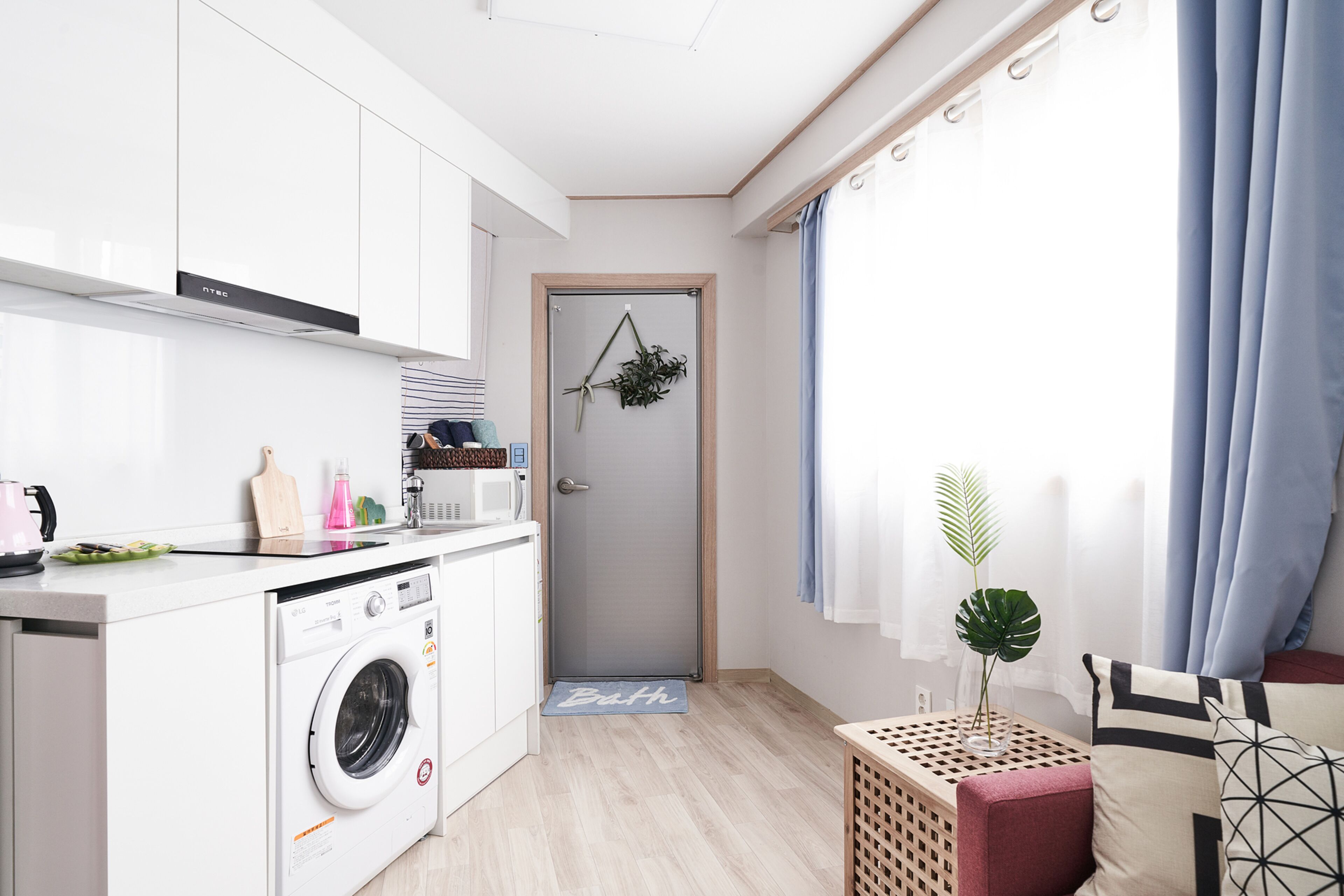 A modern kitchen with a built-in washing machine, a microwave, and a coffee maker. There is a door with a plant hanging on it, and a small table with a plant on it.