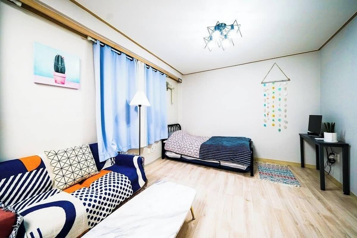 A small, well-lit room with a bed, a sofa, a desk, and a hanging light fixture.
