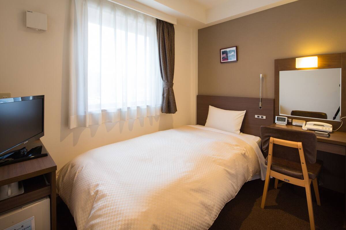 A hotel room with a bed, a desk with a chair, a television, a bedside table, and a window with curtains.