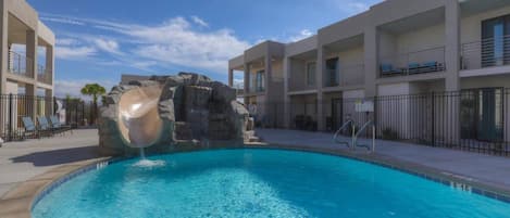 A swimming pool with a rock feature, adjacent to a building with balconies and a clear sky.