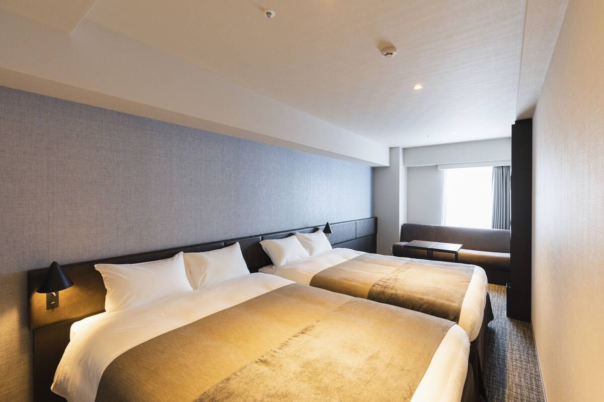 Ein Hotelzimmer mit zwei Betten, einem Nachttisch, einem Schreibtisch und einem Fenster mit Vorhängen.