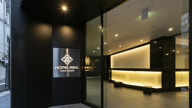 Un hall d’hôtel moderne avec une entrée en verre, un comptoir d’accueil orné d’un mur aux motifs géométriques et une enseigne indiquant « HÔTEL ASYL TOKYO HAMAMATTA ».