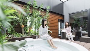 Deux femmes sont assises dans un jacuzzi extérieur, avec un bâtiment moderne et des plantes en arrière-plan.