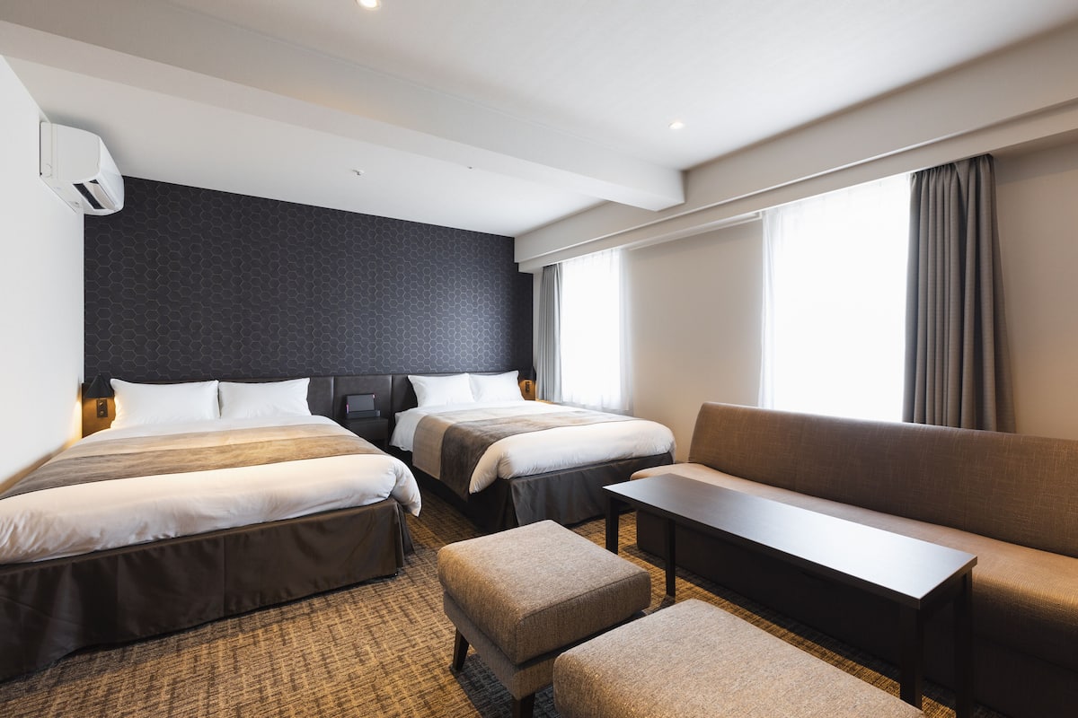 Ein Hotelzimmer mit zwei Betten, einem Schlafsofa, einem Tisch und einem großen Fenster mit Vorhängen.