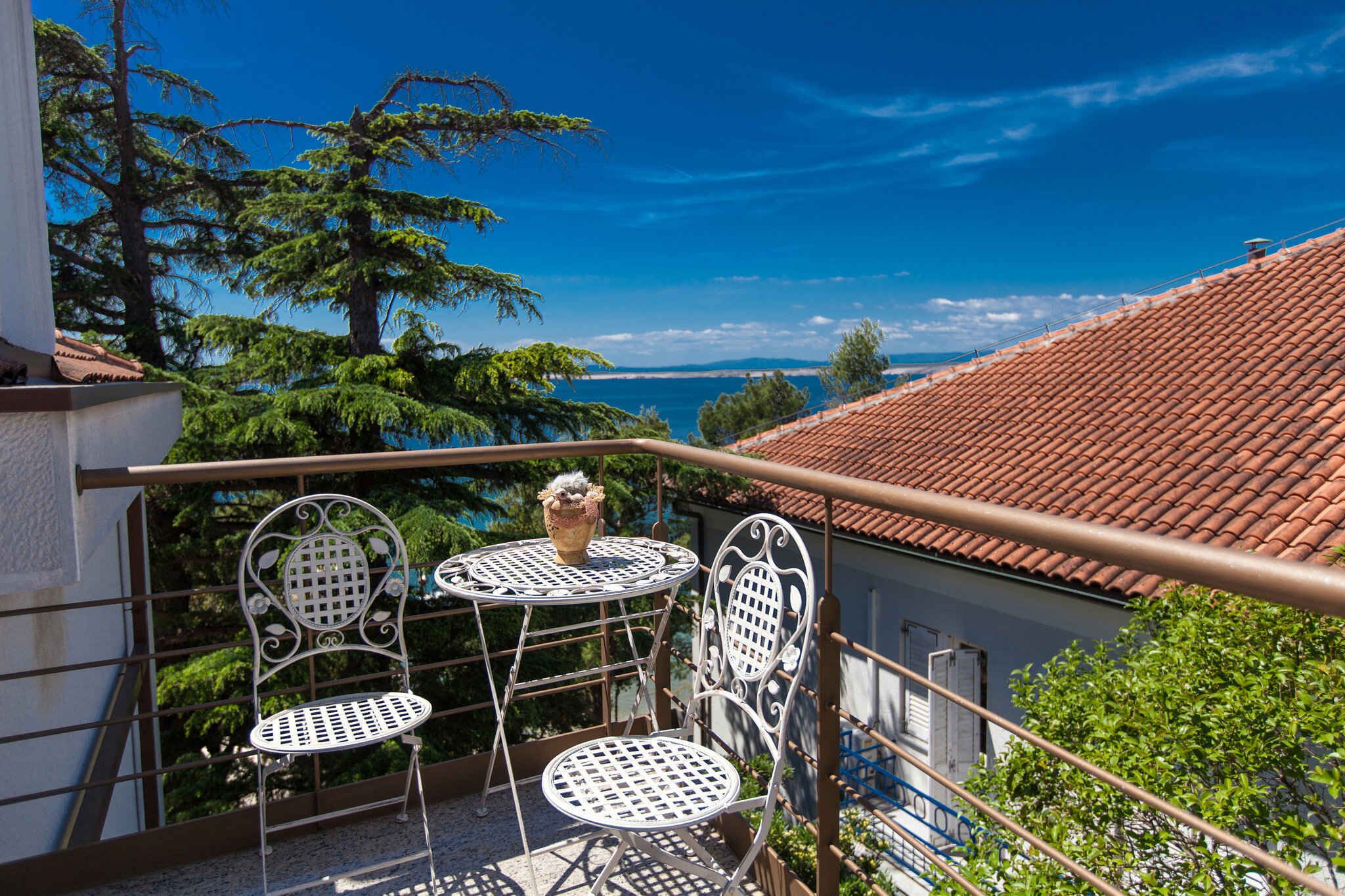 A balcony with two white metal chairs and a small table, overlooking a coastal view with trees and a red-tiled roof.