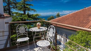 Un balcone con due sedie in metallo bianco e un tavolino, con vista sulla costa, alberi e un tetto in tegole rosse.