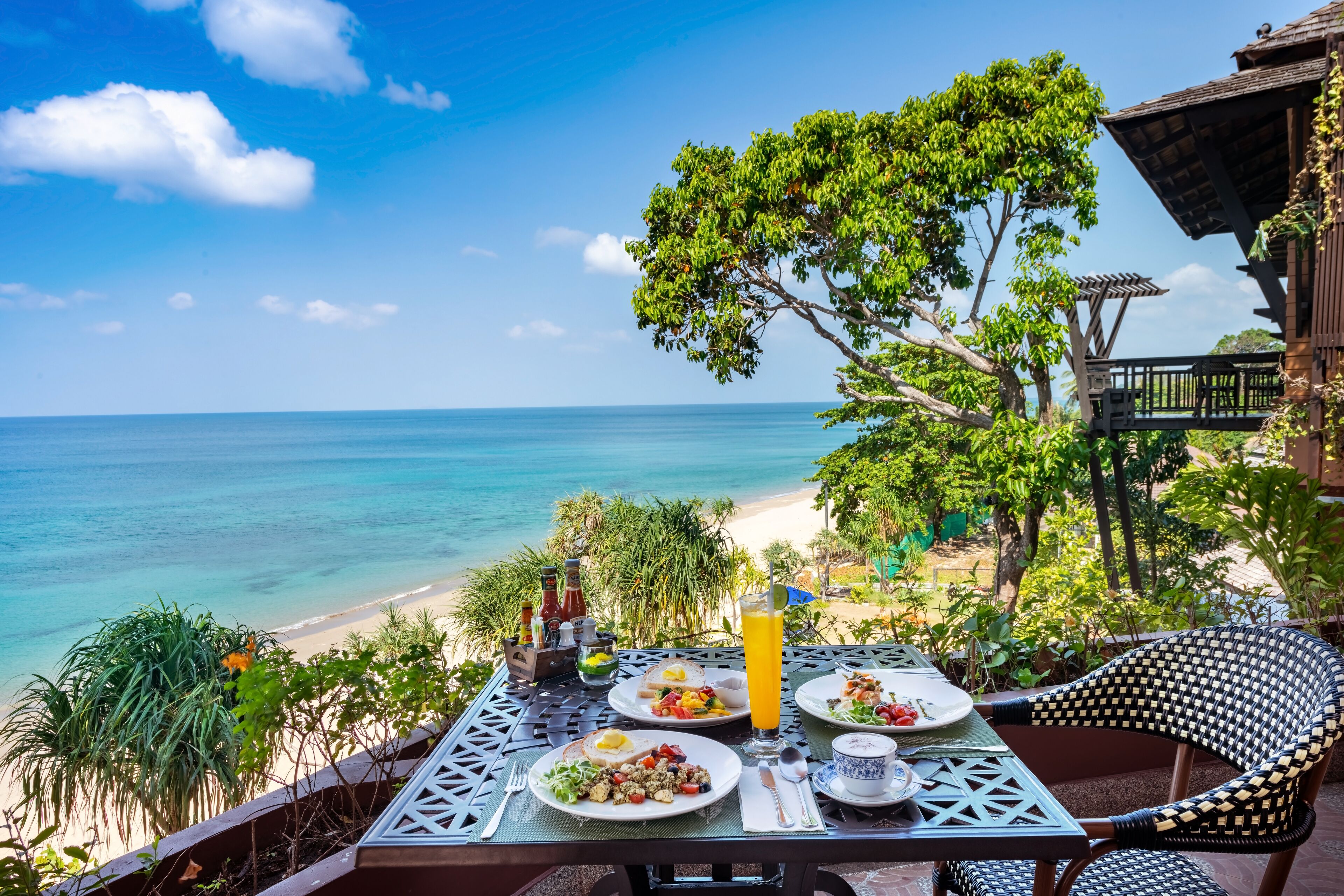 Mesa con platos de comida, un vaso de jugo y condimentos, con vista a una playa y al océano.