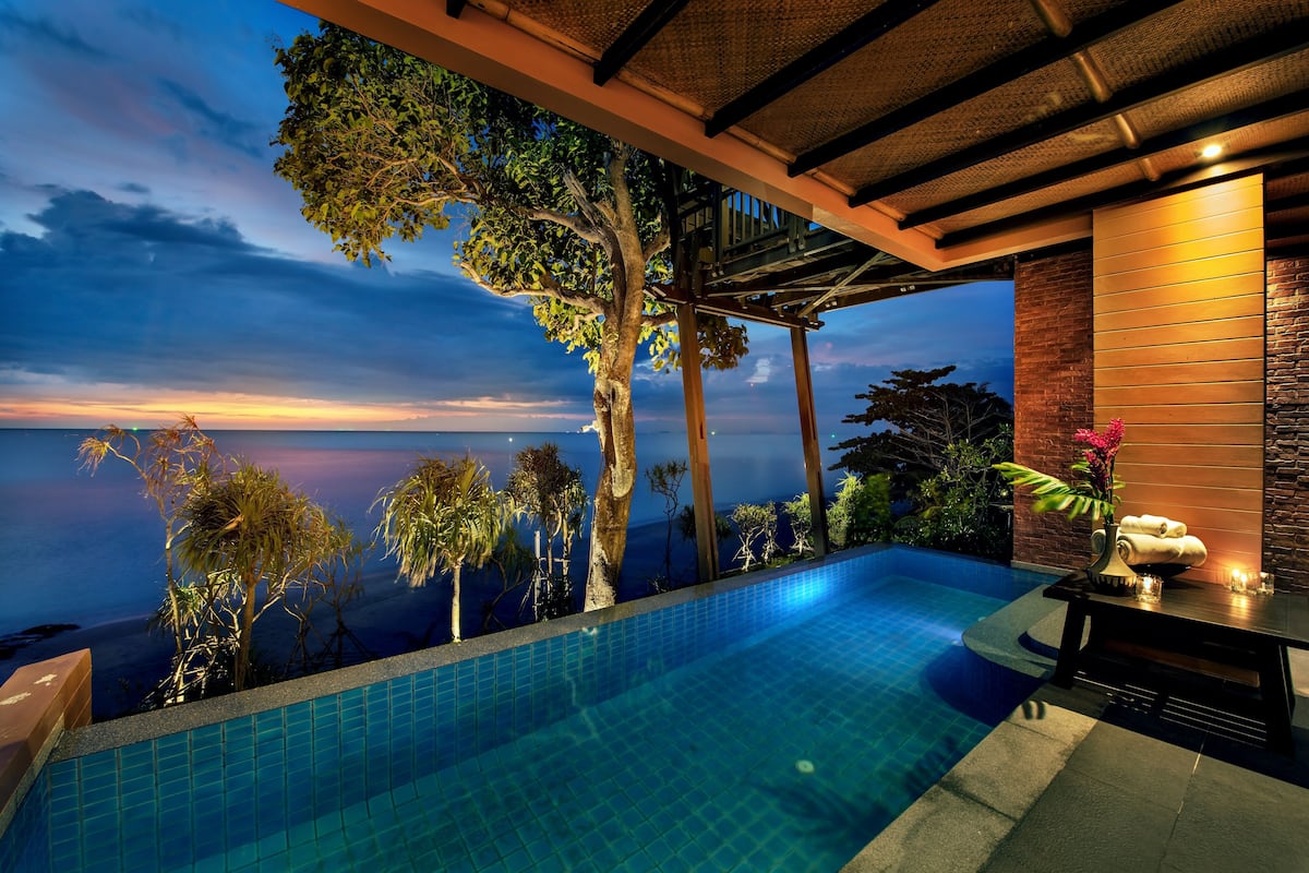 A swimming pool with a view of the ocean at dusk, surrounded by trees and a wooden deck.