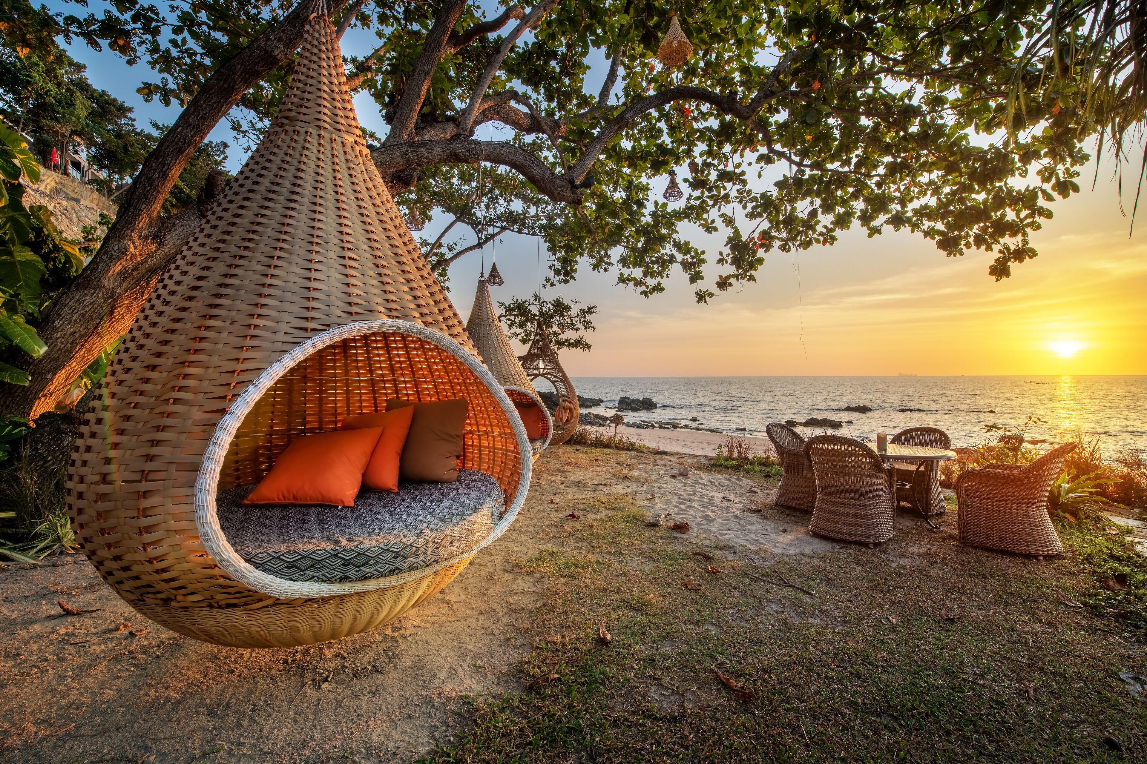 A beach setting with a hanging wicker chair and a table with chairs, overlooking the sea.