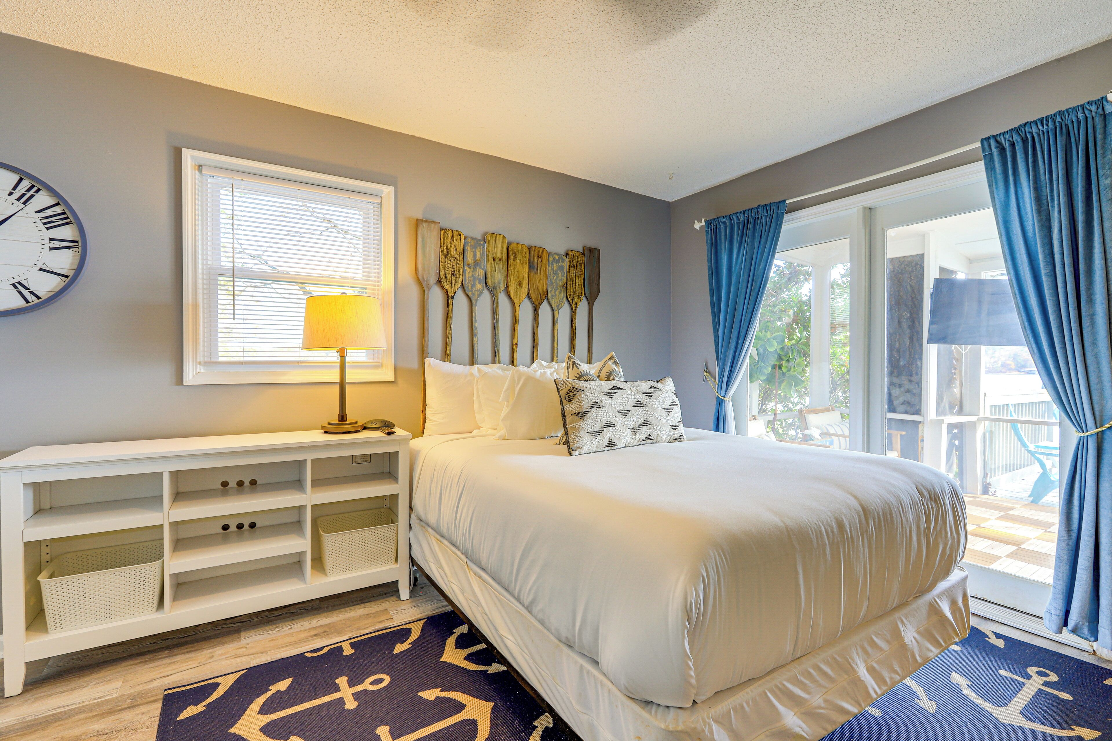 A bedroom with a bed, a nightstand, a lamp, a wall clock, and a window with curtains.