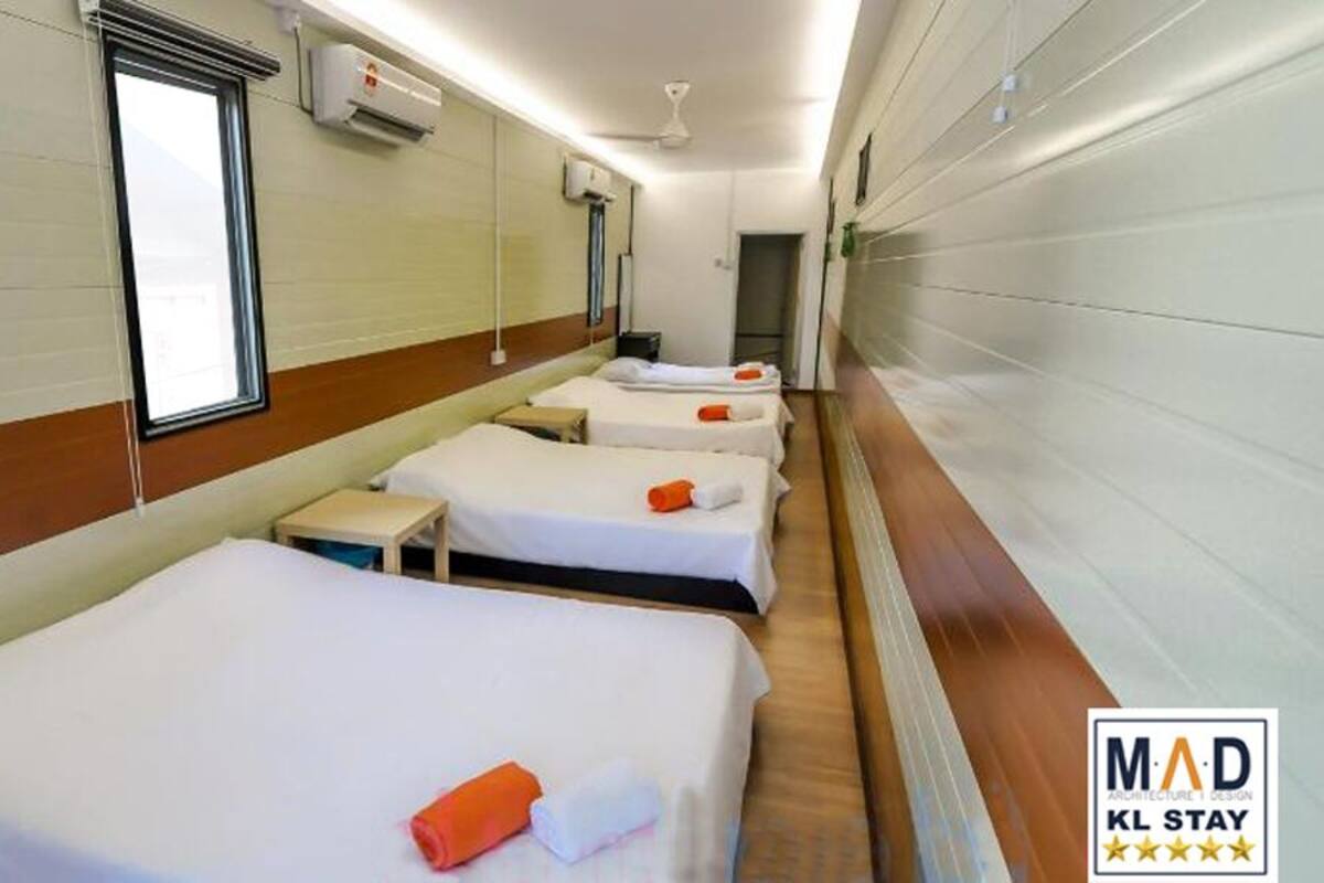 Ein Hotelzimmer mit drei Einzelbetten, jedes mit einer weißen Matratze und orangefarbenen Handtüchern. An den Wänden befinden sich Klimaanlagen, ein Deckenventilator und ein Fenster, das für natürliches Licht sorgt.