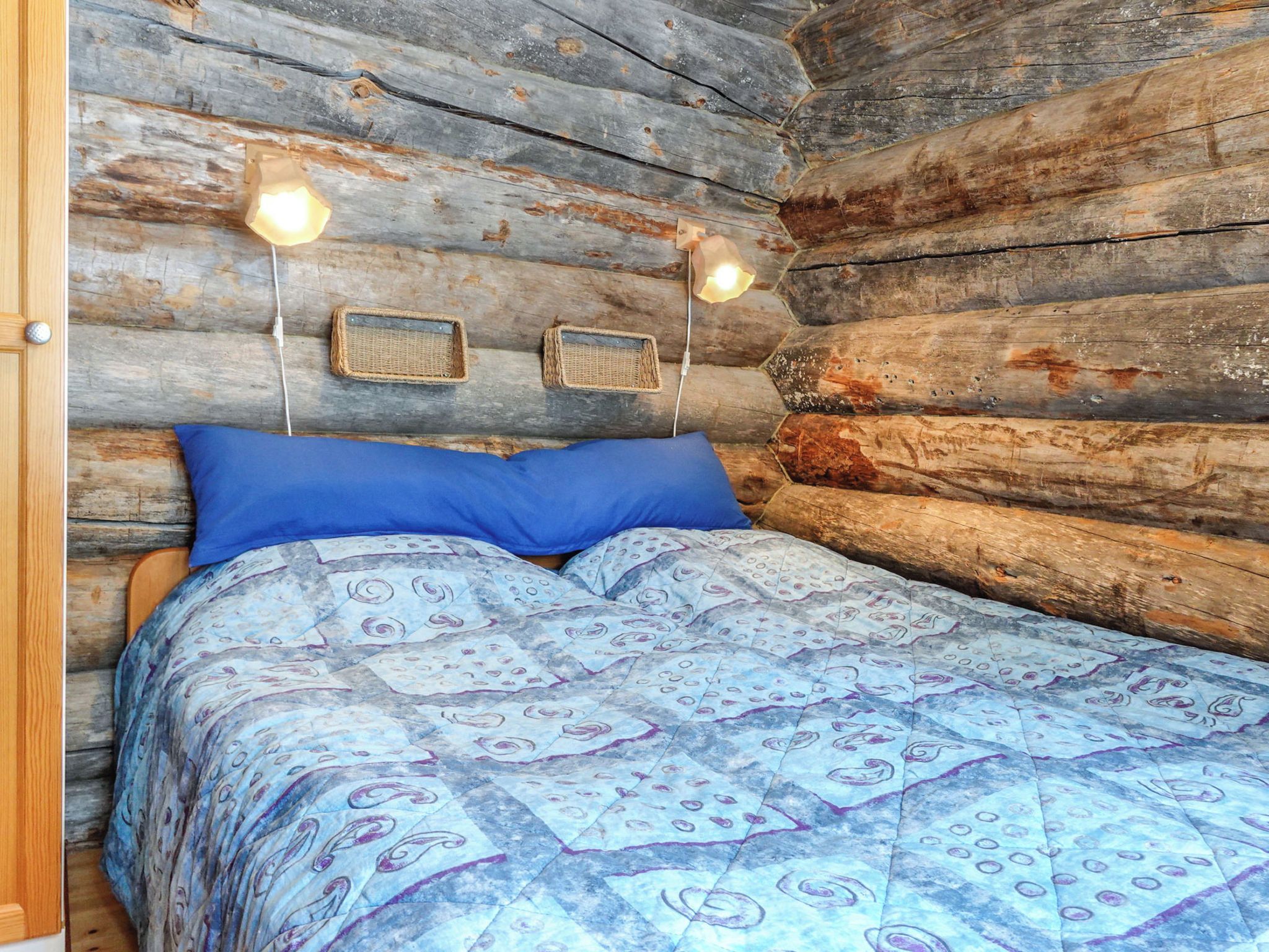 Interior d'una cabana de fusta amb un llit, dues làmpades penjades a la paret i una porta.