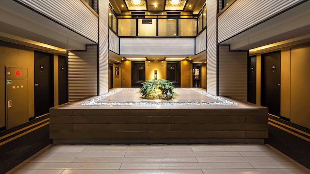 A modern hotel lobby with a central water feature, multiple elevators, and a glass-enclosed upper level. - APA Hotel Ueno Hirokoji (Tokyo)