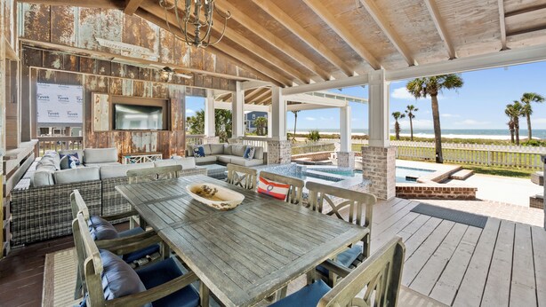 Ein überdachter Außenbereich mit Holztisch und Bänken, einem Fernseher und Blick aufs Meer.