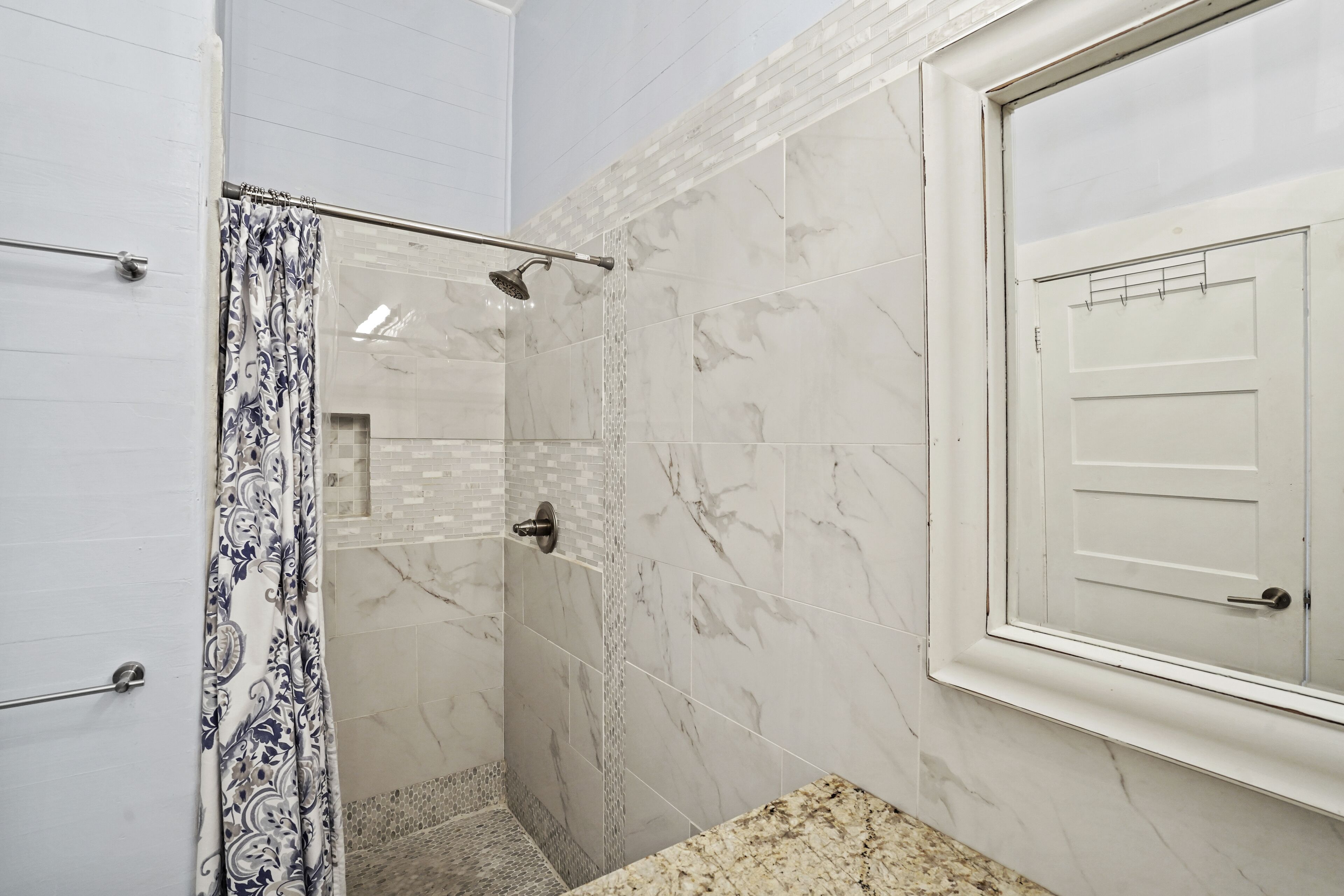 A bathroom with a shower, marble walls, a patterned curtain, and a door with a window.