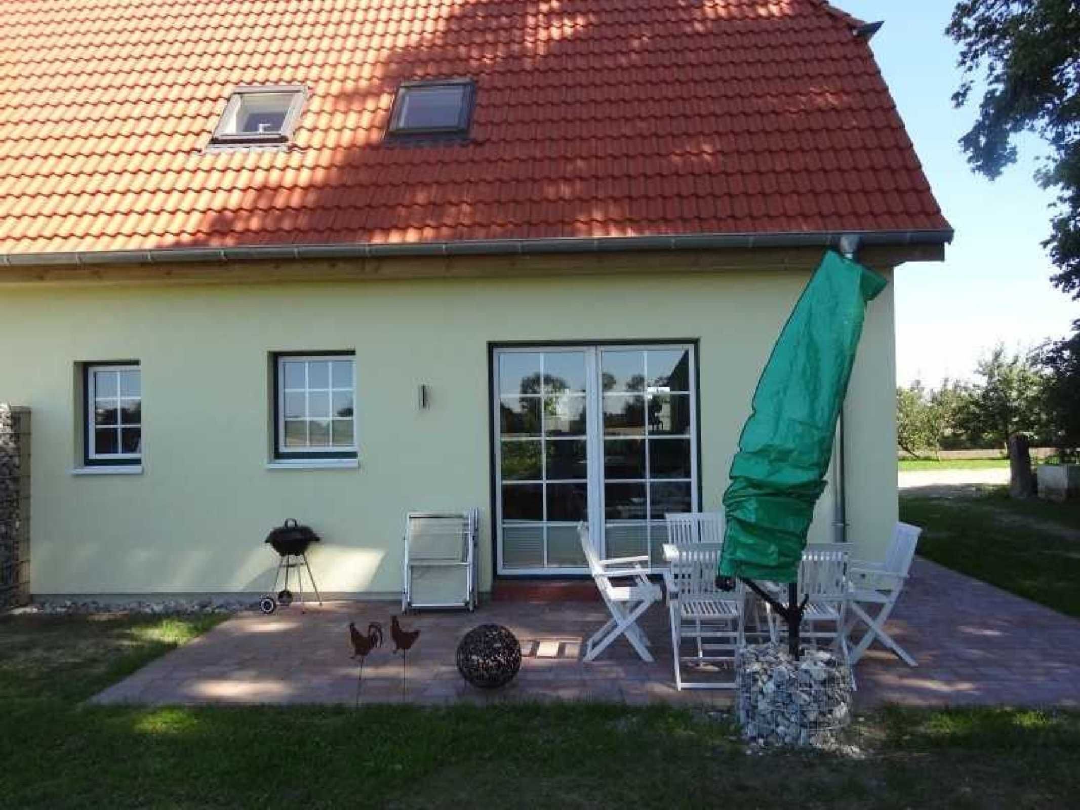 Uma casa com telhado de telhas vermelhas, uma grande porta de correr de vidro e um alpendre com cadeiras brancas e churrasqueira.