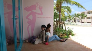 Two people sitting outside a hotel entrance with a palm tree and a building in the background.
