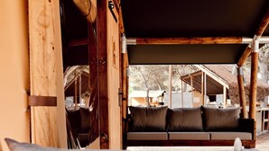 A wooden interior with a bench and cushions, a wooden door, and a view of an outdoor area with a building and trees.