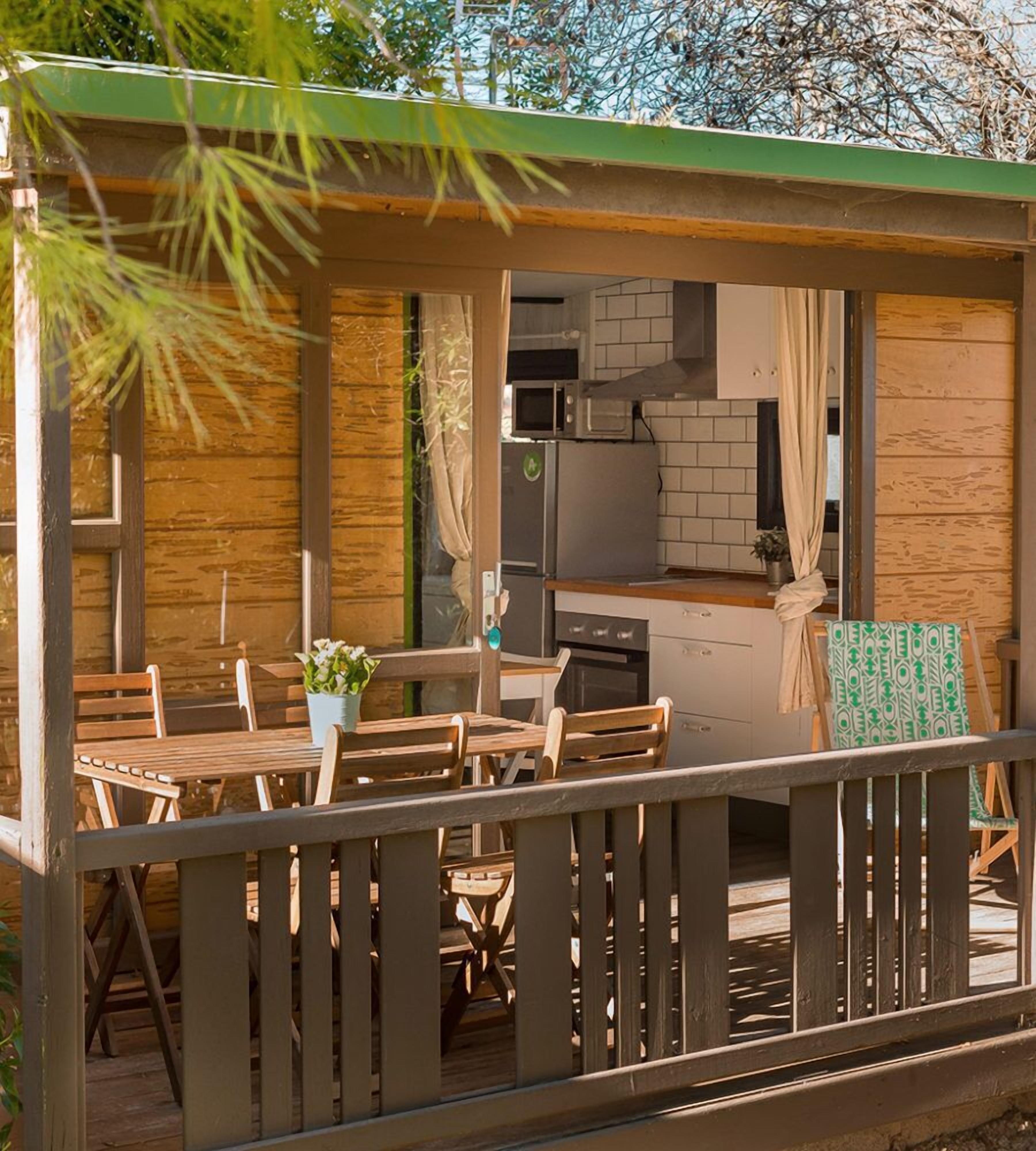 Una terraza de madera con una mesa y sillas, una pequeña cocina equipada con microondas y refrigerador, y vistas a árboles y al cielo.