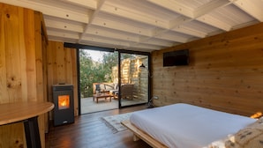 A wooden cabin room with a bed, a table, a fireplace, and a sliding glass door leading to an outdoor seating area.