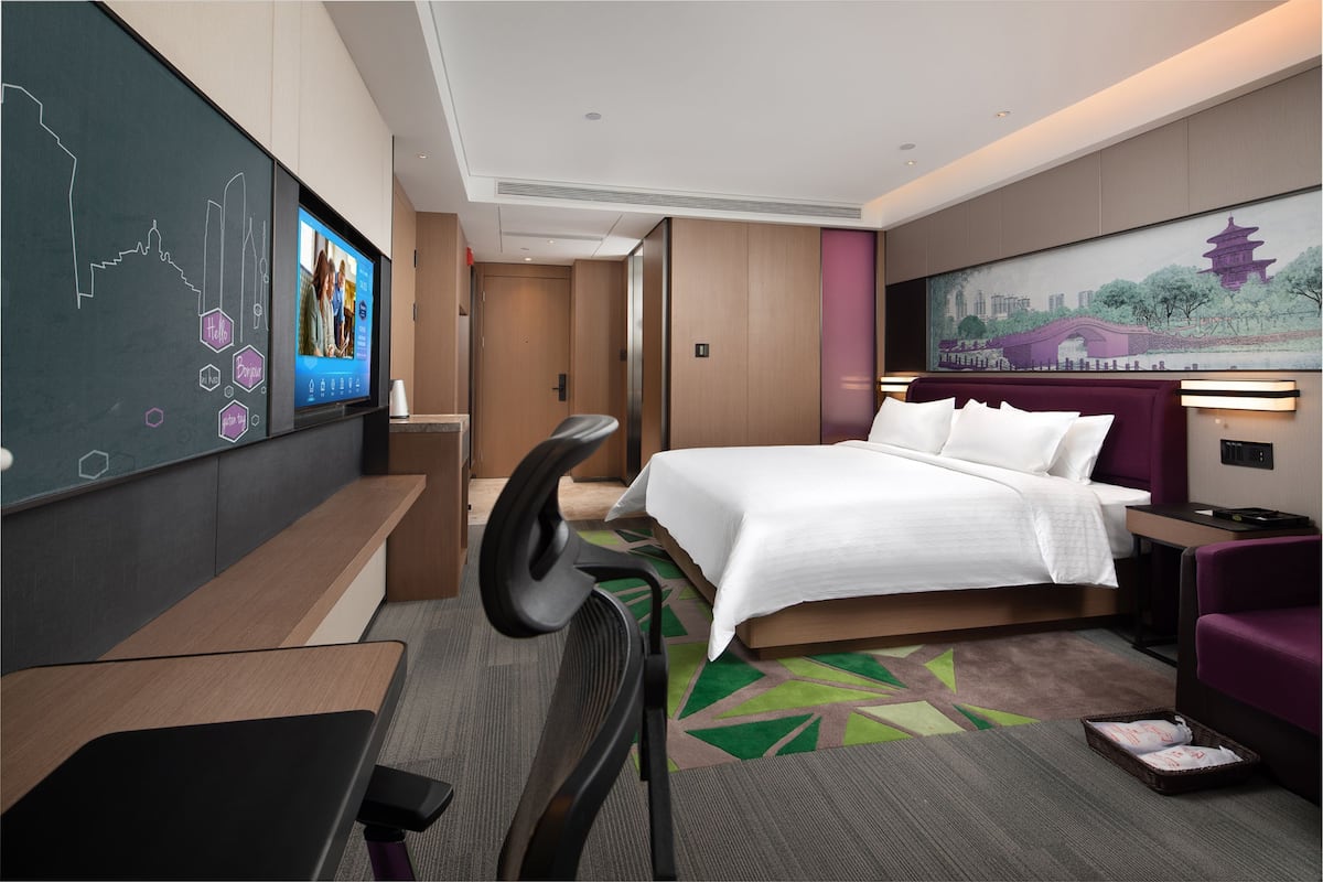 A hotel room with a bed, a desk, a chair, a TV screen, and a painting on the wall.
