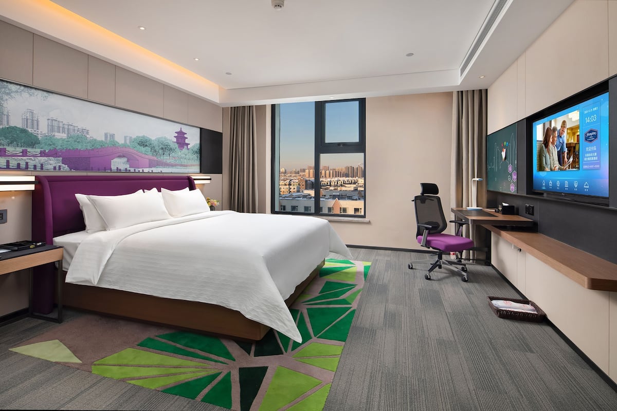 A hotel room with a large bed, a desk with a computer, a television, and a city view.