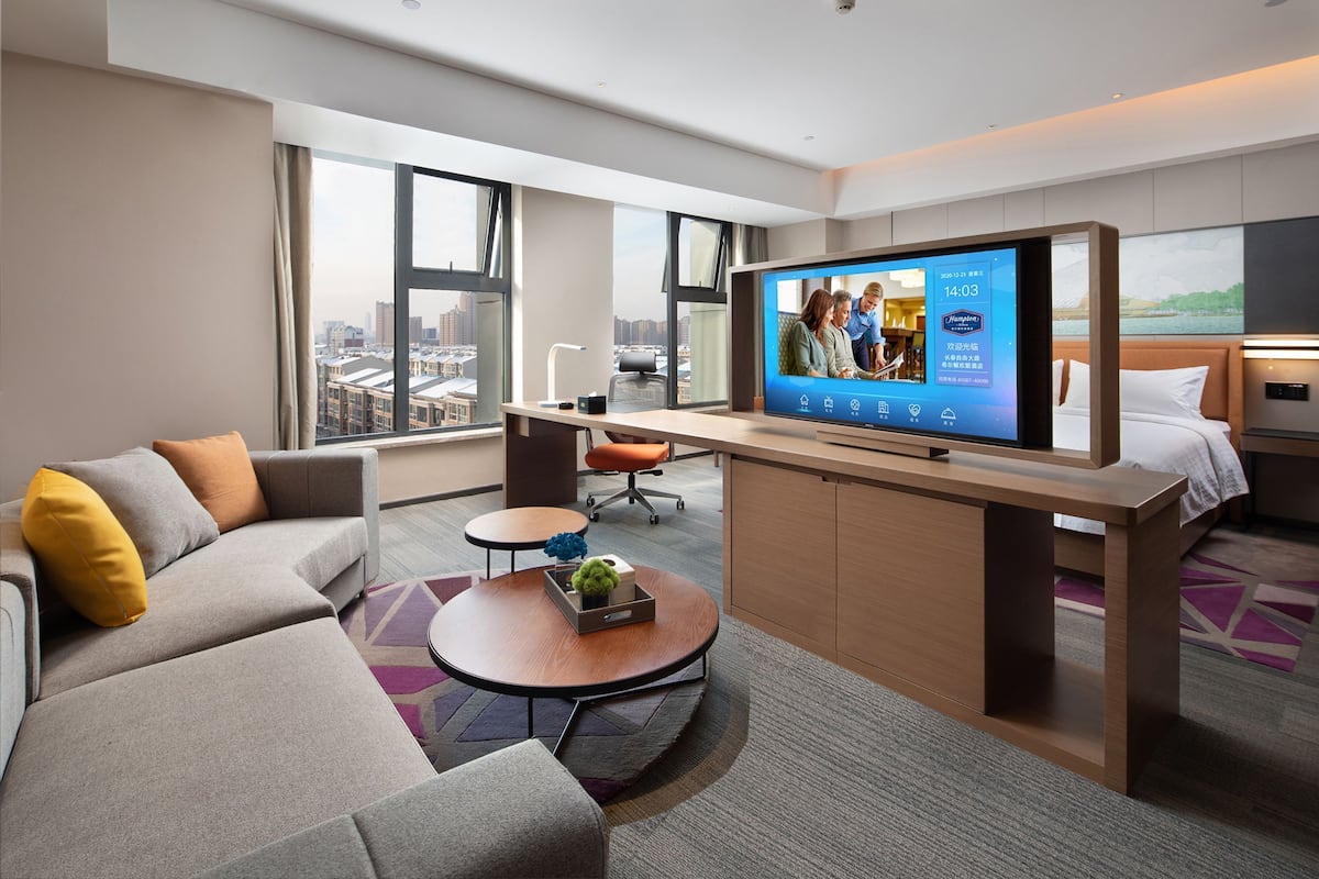A modern hotel room with a large TV, a sofa, a desk, and a bed.