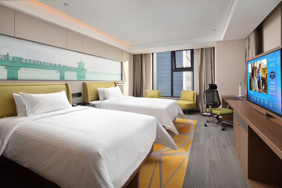 A hotel room with two beds, a desk with a TV, a chair, and a large window.