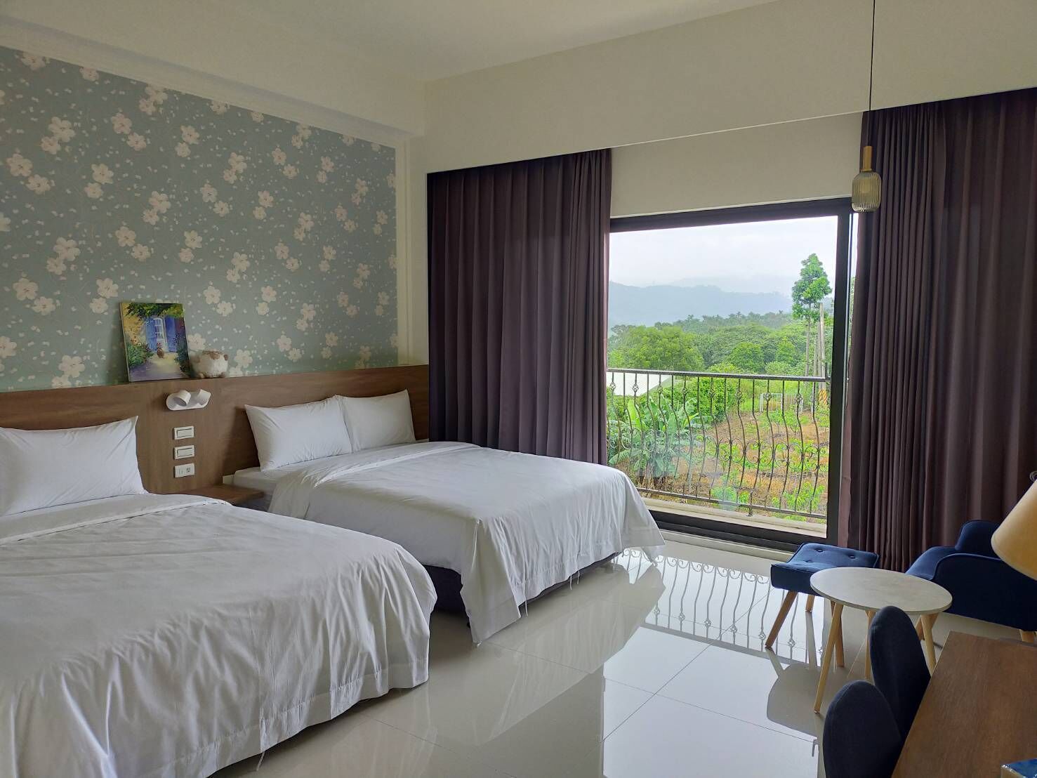 Habitación de hotel con dos camas, un balcón con vistas a zonas verdes y un pequeño rincón de descanso con una mesa y sillas.
