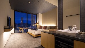 Une chambre d’hôtel moderne équipée d’un lit, d’un bureau, d’une grande fenêtre donnant sur la ville, d’une salle de bain avec un lavabo et un miroir, et d’un téléviseur placé à gauche.