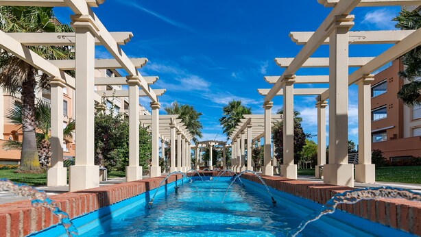Una piscina con agua cristalina azul, rodeada de columnas blancas y palmeras.