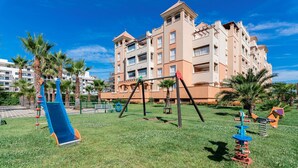 Un parque infantil con tobogán y columpios, rodeado de palmeras y un edificio con balcones.