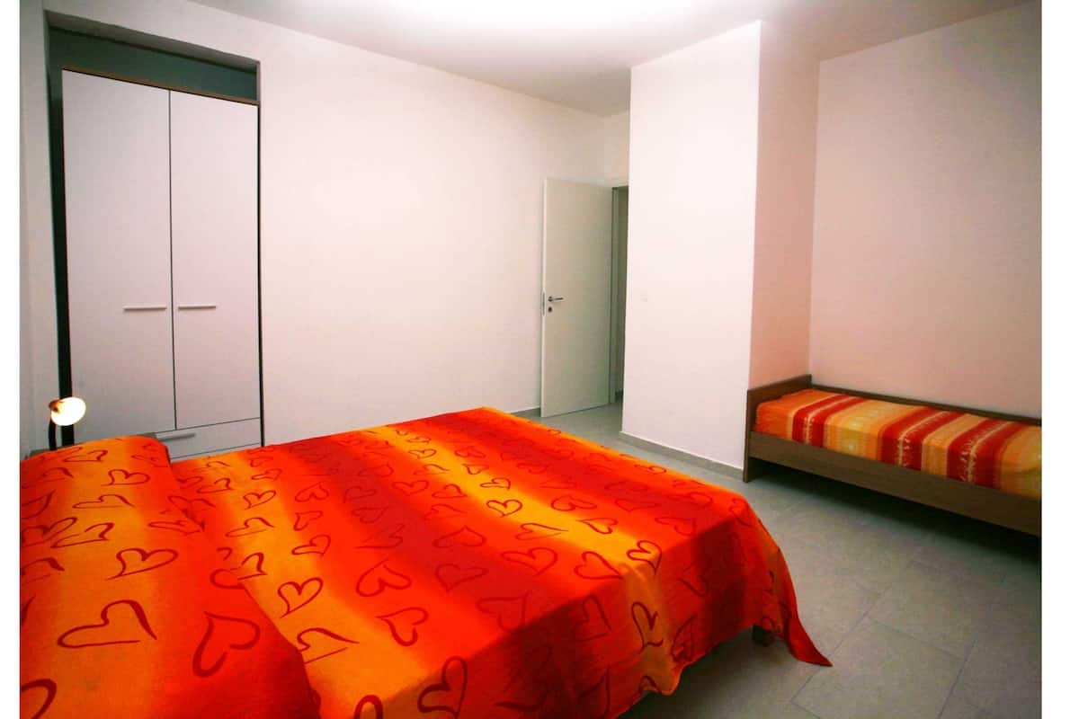 Une chambre à coucher avec un lit recouvert d’une couverture rouge à motifs en forme de cœur, un banc en bois avec un coussin rayé et une armoire.