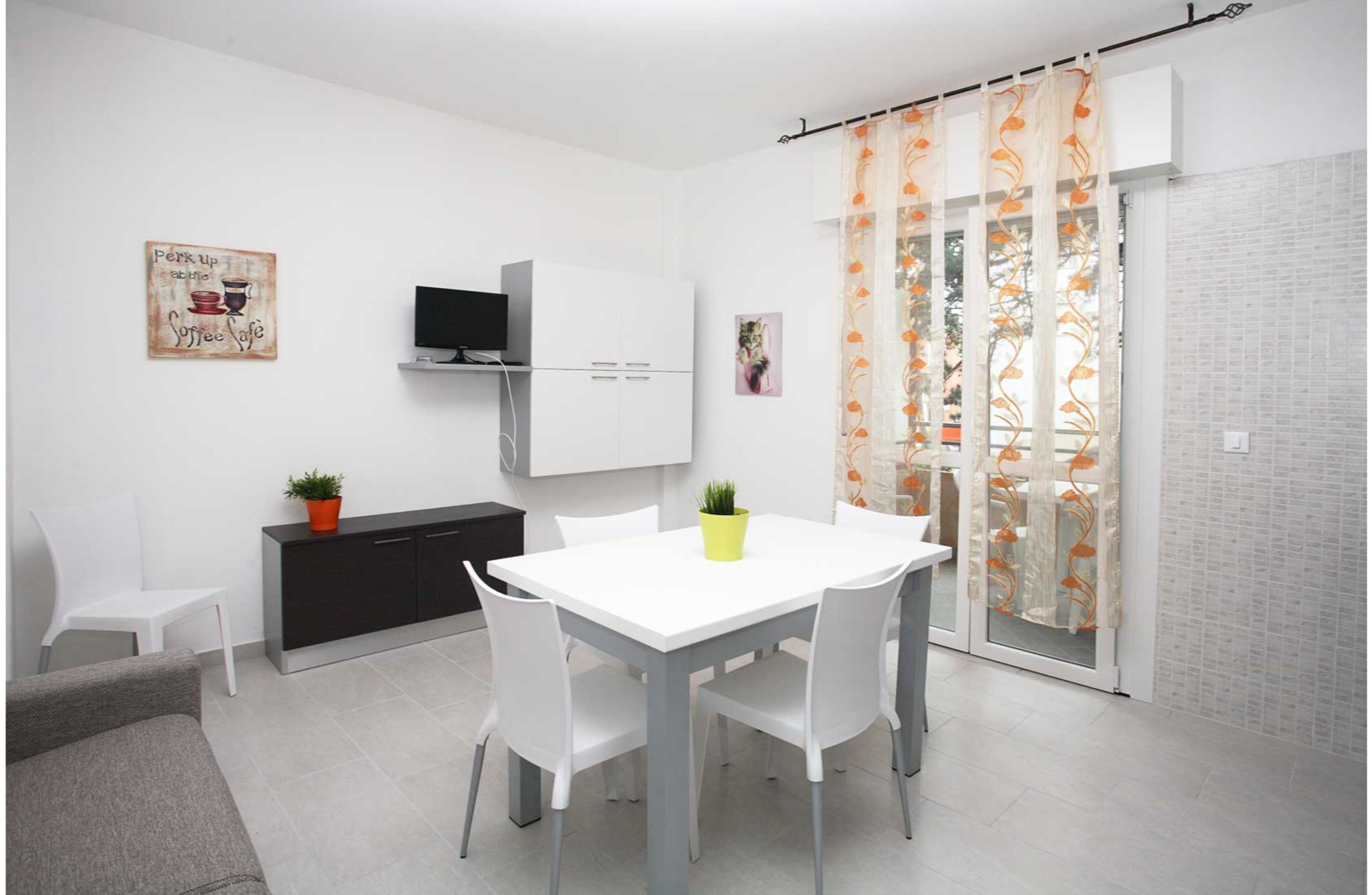 A modern, well-lit room with a white dining table, two chairs, a small black cabinet, a TV mounted on the wall, and a balcony with a patterned curtain.
