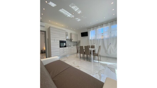 A modern living room with a sofa, a dining area with chairs, a kitchen with cabinets and a TV, and a large window with curtains. - Appartamenti Boschetto (San Michele al Tagliamento)