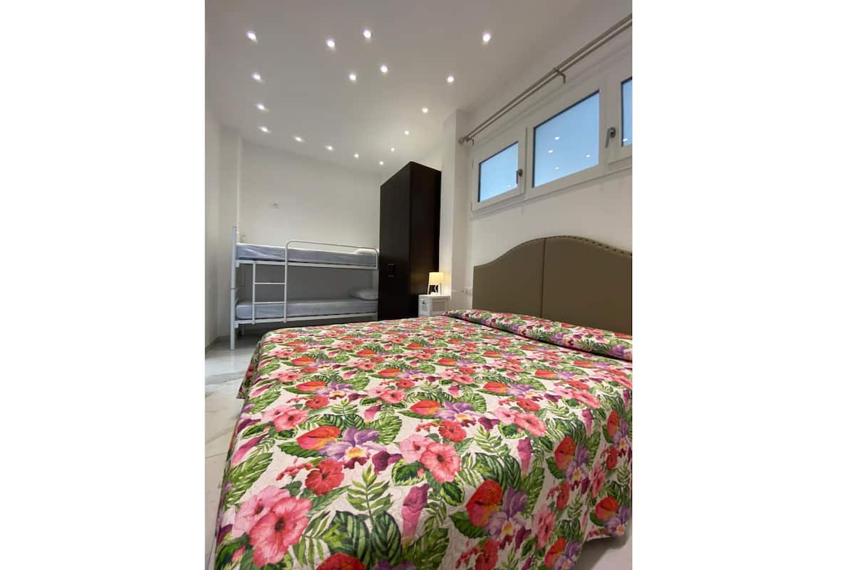 Une chambre avec un lit superposé, un grand lit recouvert d’une couvre-lit à motifs floraux, une tête de lit, une fenêtre et des spots encastrés au plafond.