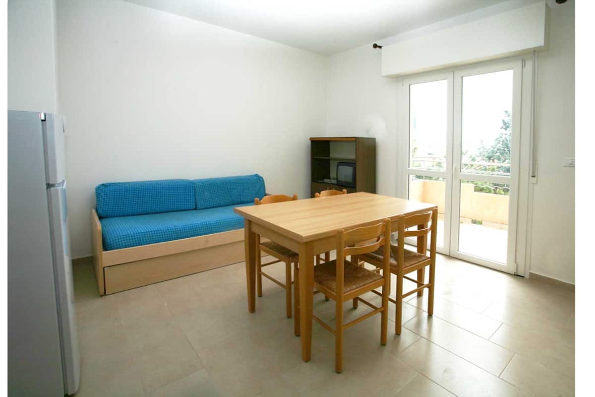 Une petite pièce lumineuse, avec une table et des chaises en bois, un canapé bleu, un téléviseur posé sur une étagère et une porte-fenêtre coulissante donnant sur un balcon.