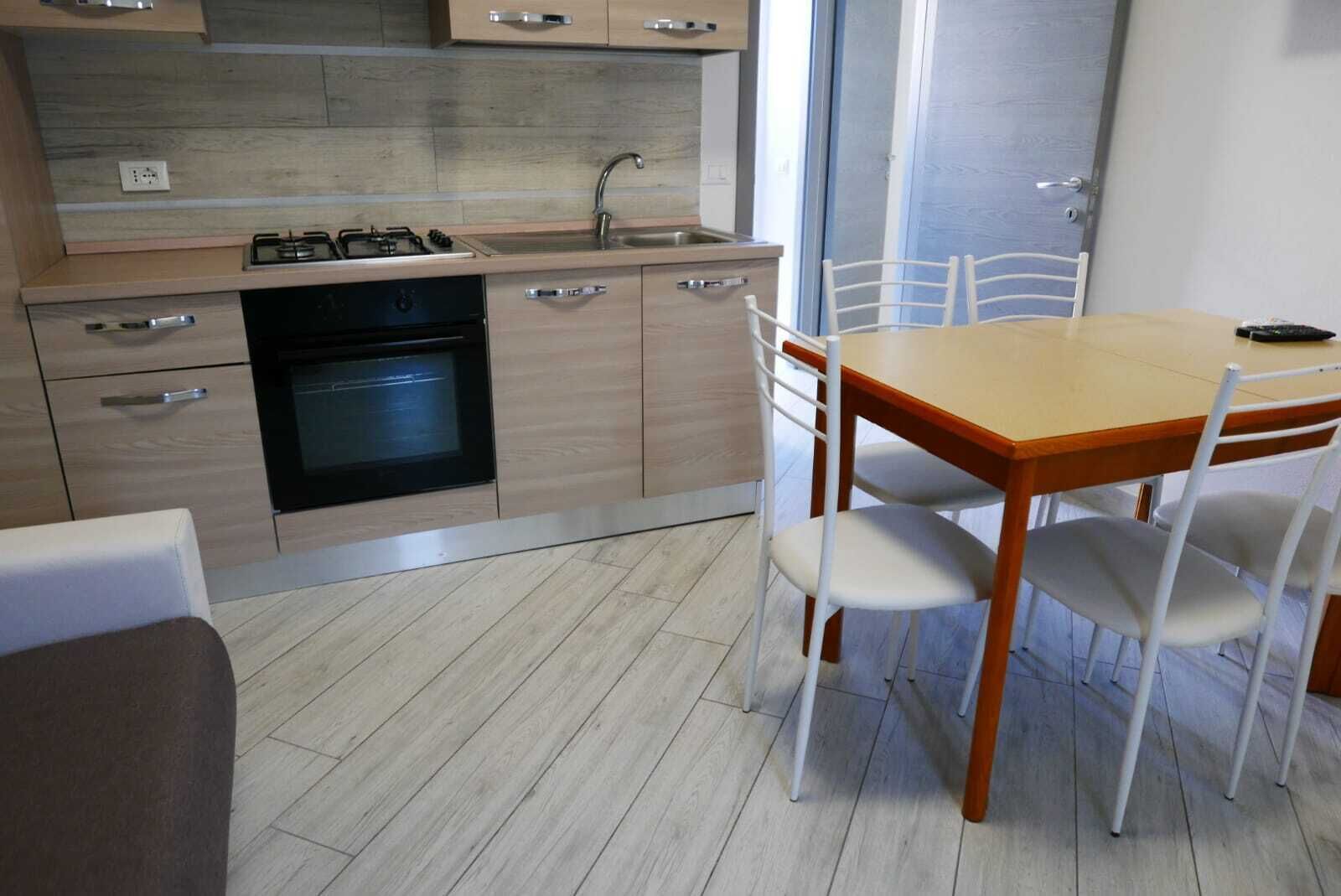 A compact kitchen with a gas stove, oven, and sink. There is a dining table with four chairs. The room has a tiled floor and light-colored walls.