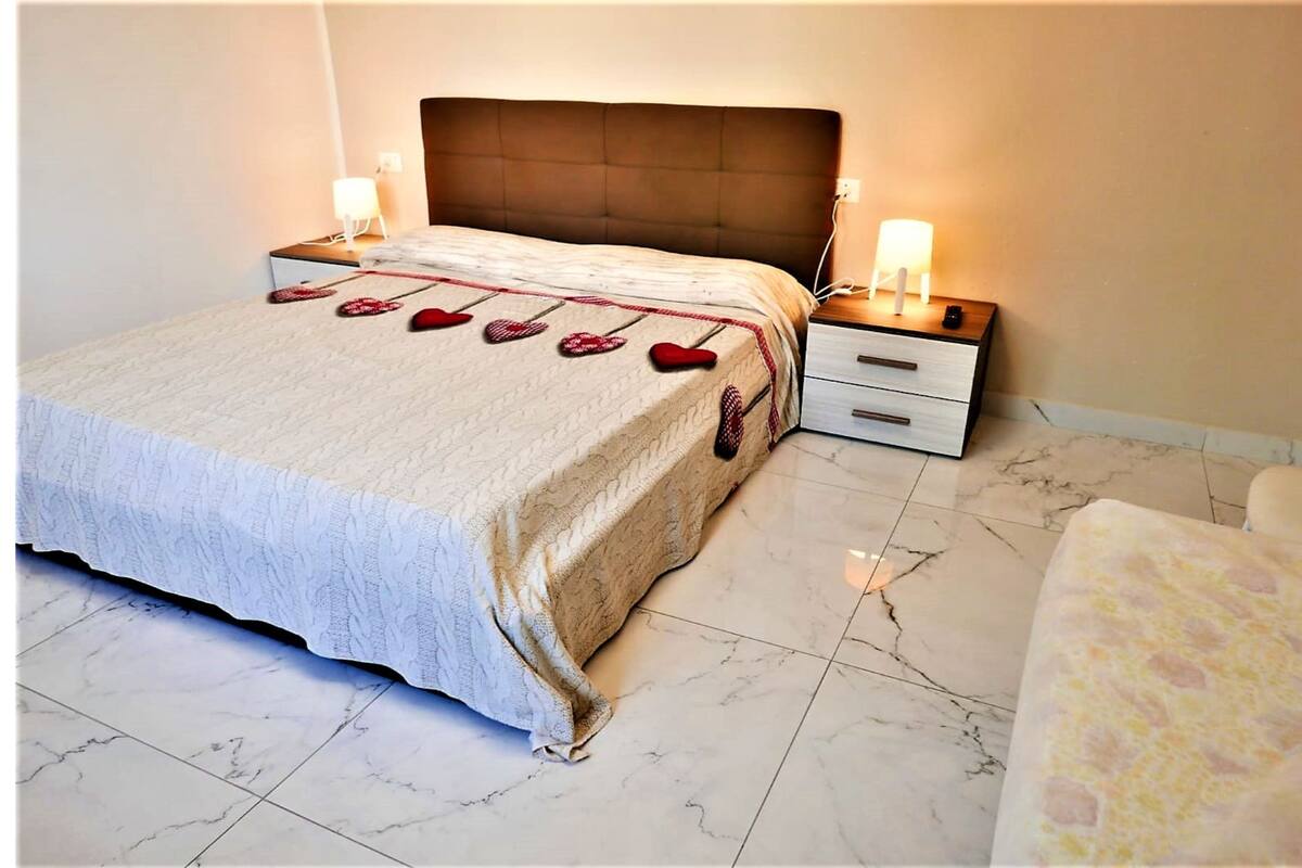 Un lit bien fait, recouvert d’une couvre-lit à motifs de cœurs, deux tables de chevet avec des lampes et un sol en marbre.