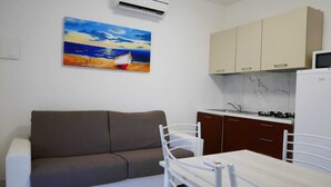 Ein kompakter Wohnbereich mit einer Couch, einem Esstisch, Stühlen, einer Küchenzeile mit Schränken und einem Kühlschrank, sowie einem Gemälde einer Strandansicht.