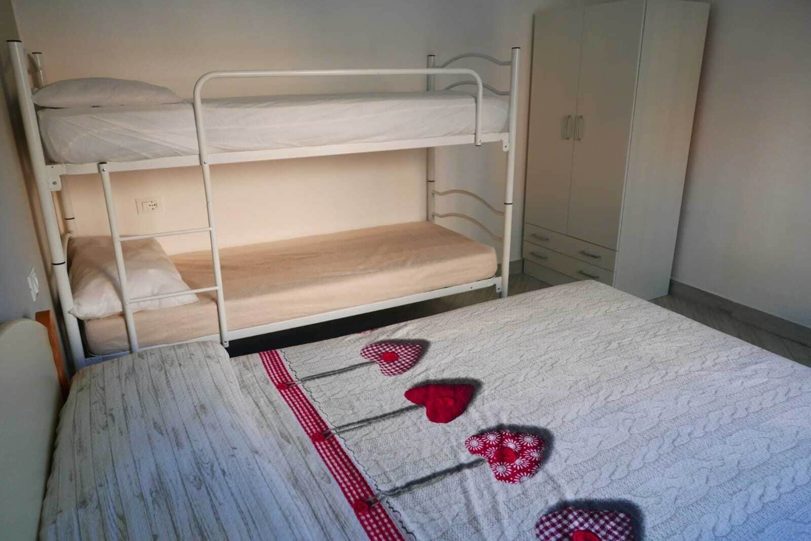Una cama litera con una cama inferior y otra individual debajo. Hay un armario a la derecha y un enchufe en la pared a la izquierda.