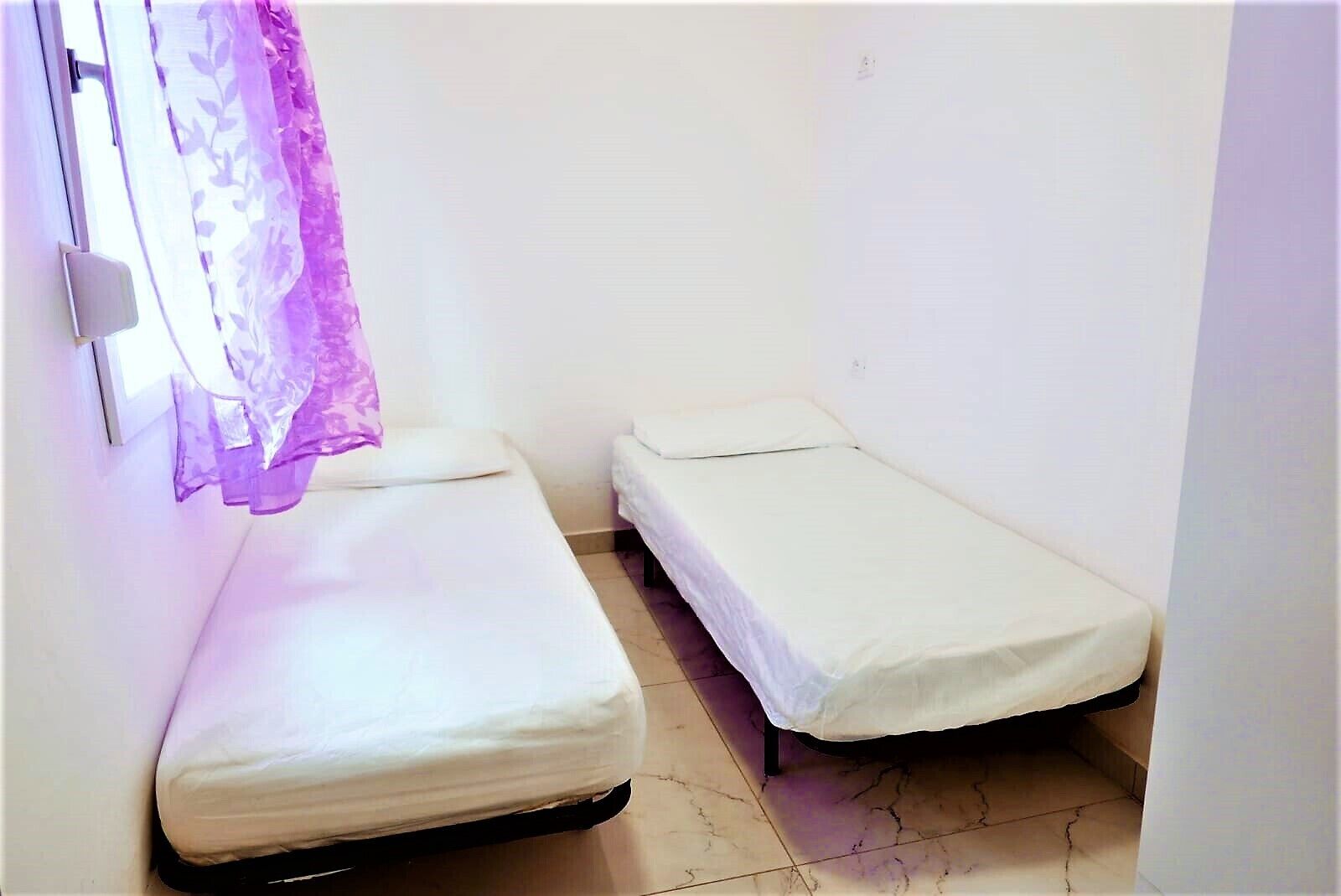 Un petit logement composé d’une seule pièce, avec deux lits simples, une fenêtre ornée d’un rideau violet et un mur blanc.