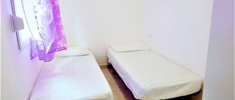 A small, single-room accommodation with two single beds, a window with a purple curtain, and a white wall.