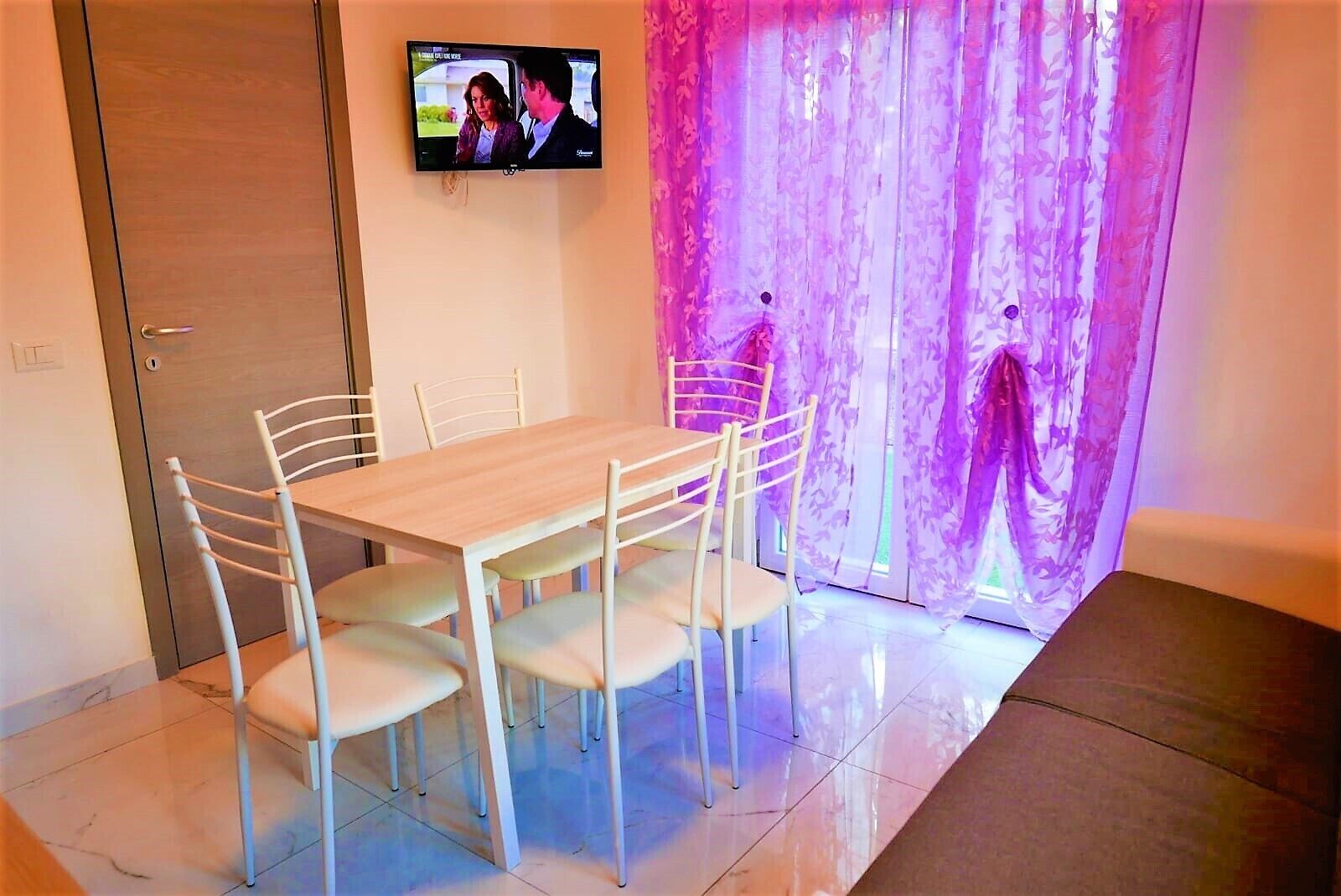 Un comedor con una mesa de madera y sillas blancas, un televisor montado en la pared y una cortina morada con estampado floral.
