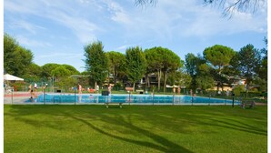 A swimming pool surrounded by a fence, with trees and a grassy area in the foreground. - Appartamenti Olimpia (San Michele al Tagliamento)