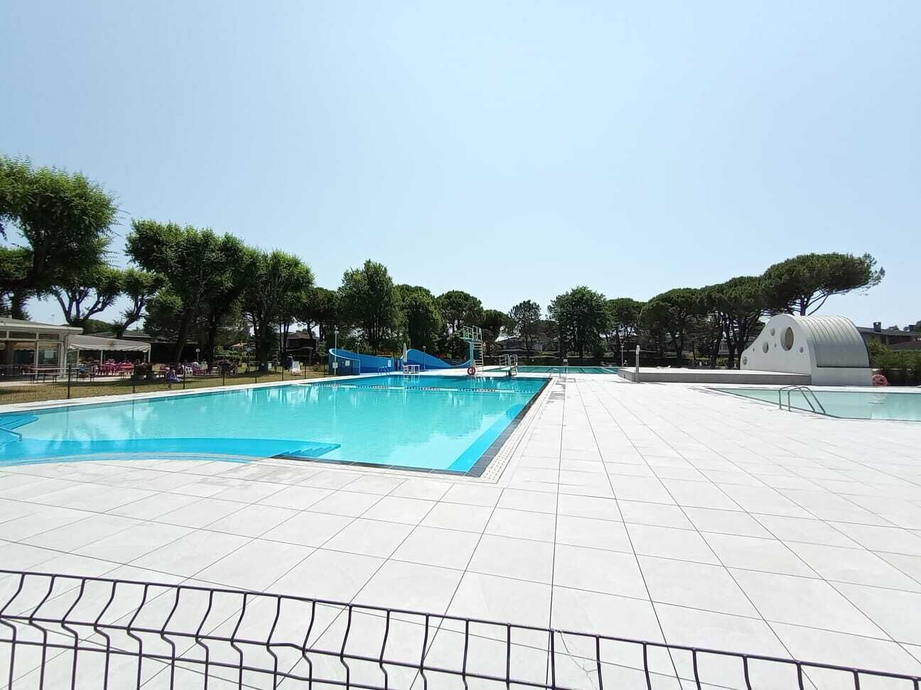 A swimming pool with a tiled deck, surrounded by trees and a building with a dome-shaped structure.